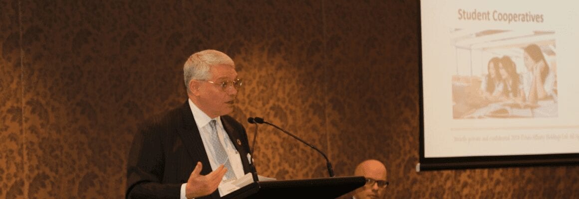 An older man in a suit speaks at a podium in a conference room. A presentation slide titled Student Cooperatives with a group photo is projected on the screen beside him. Another person sits in the background.