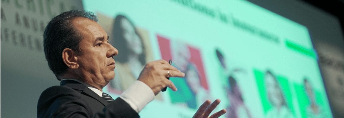 A man in a suit gestures while speaking on stage in front of a large screen displaying blurred images and text.