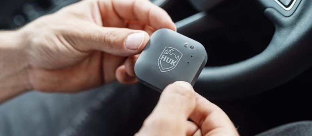 A person holds a small, square, gray electronic device with the HUK logo, inside a car near the steering wheel.