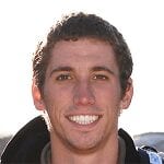 A young man with short, curly brown hair is smiling at the camera. He is outdoors with bright sunlight behind him, wearing a dark jacket and carrying a backpack.