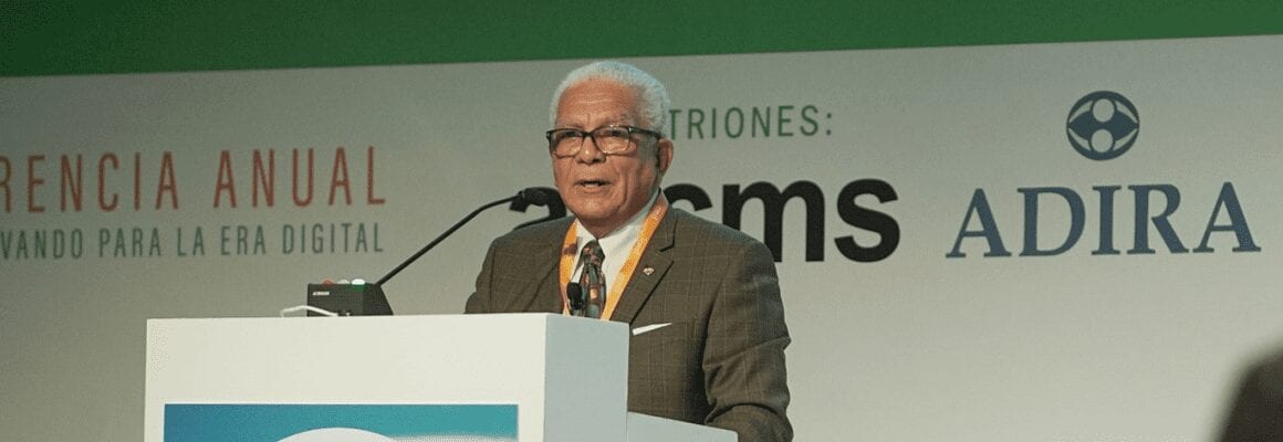 An older man with white hair and glasses speaks at a podium during a conference. Behind him are banners with the logos asms and ADIRA and part of a sign in Spanish. He wears a suit and an orange lanyard.