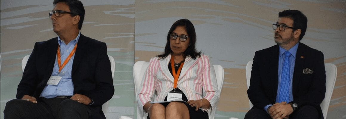 Three people wearing business attire, orange lanyards, and name tags are seated on a stage, facing forward. The background is neutral with a subtle abstract design.