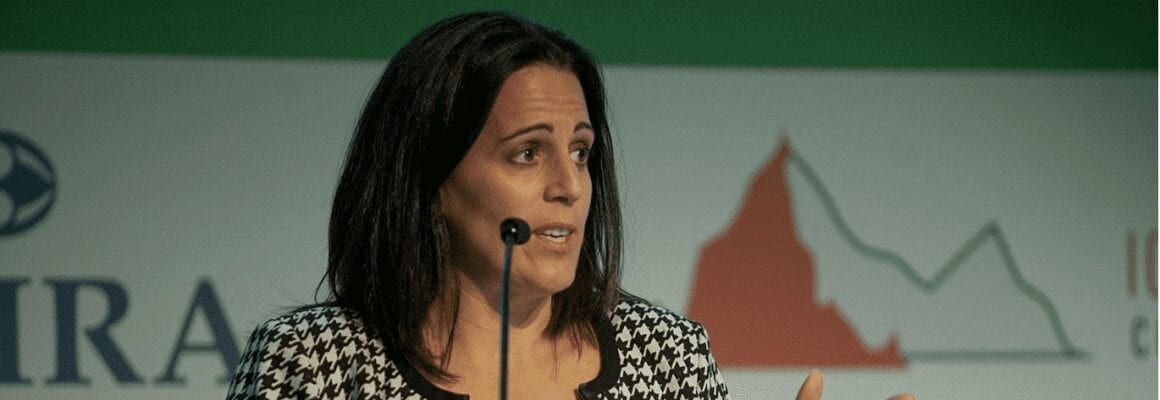A woman with dark hair, wearing a black and white patterned jacket, speaks at a podium. Behind her is a backdrop with logos and a red and green mountain graphic.