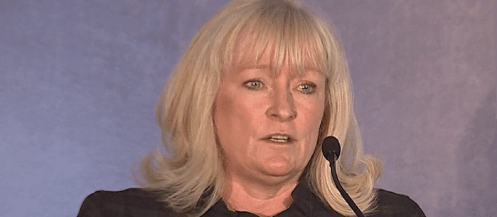 A woman with blonde hair and bangs speaks into a microphone at a podium, wearing a dark outfit, against a plain purple-grey background.