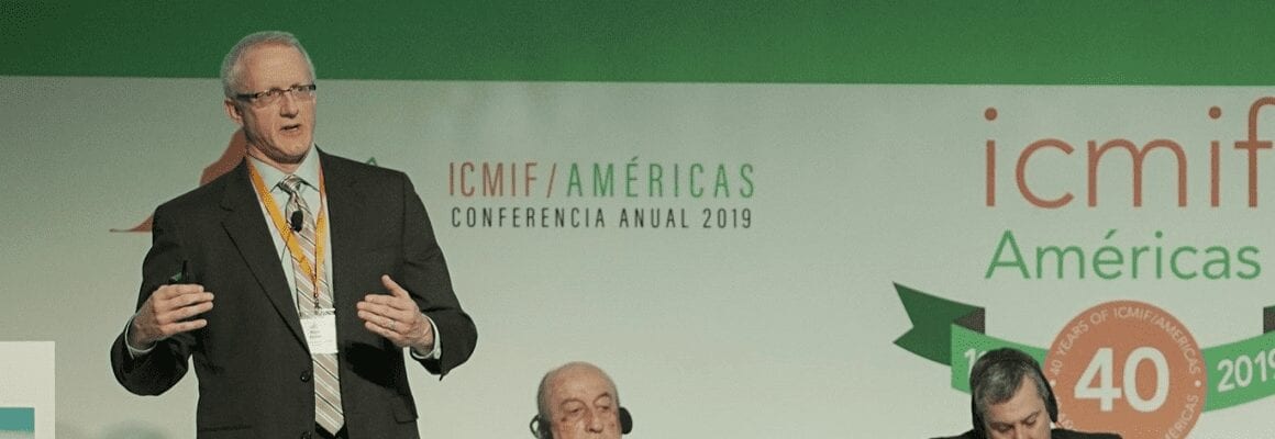 A man in a suit speaks on stage at the ICMIF Americas Annual Conference 2019, with two other men seated behind him and a conference banner in the background.