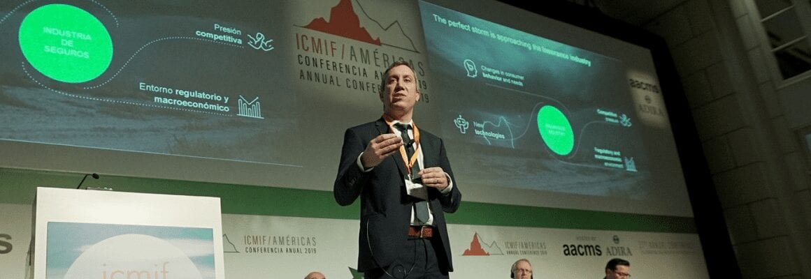 A man in a suit speaks on stage at a conference. Behind him, a large screen displays graphs and text about the insurance industry. Three seated panelists are visible in the background.