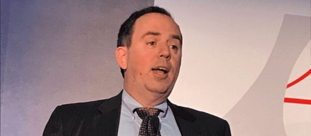 A man in a suit and tie is speaking in front of a light-colored background with abstract shapes. He appears to be giving a presentation or speech.