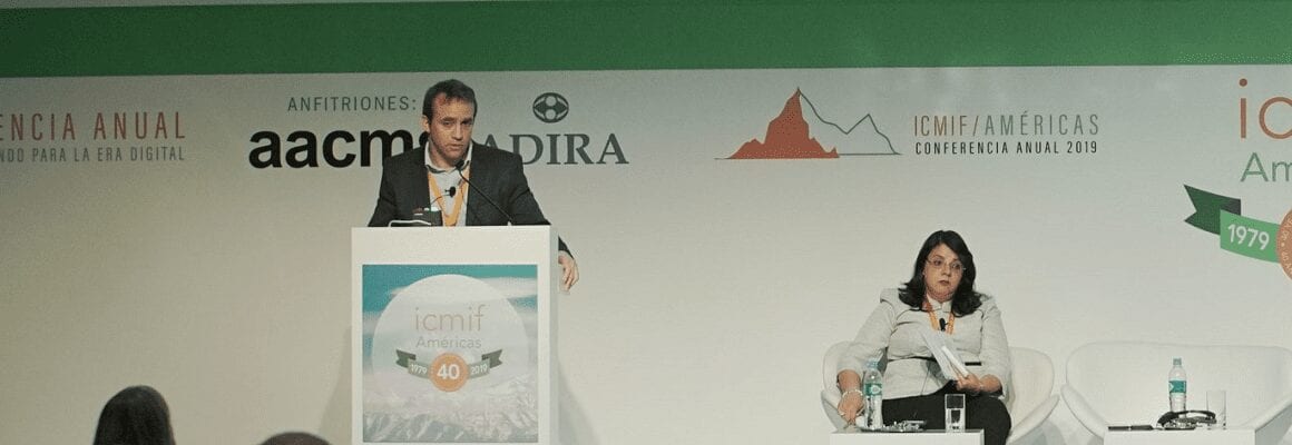 A man stands at a podium speaking while a woman sits on a white chair with a microphone in hand during the ICMIF Americas Annual Conference 2019. Event banners and logos are displayed behind them.
