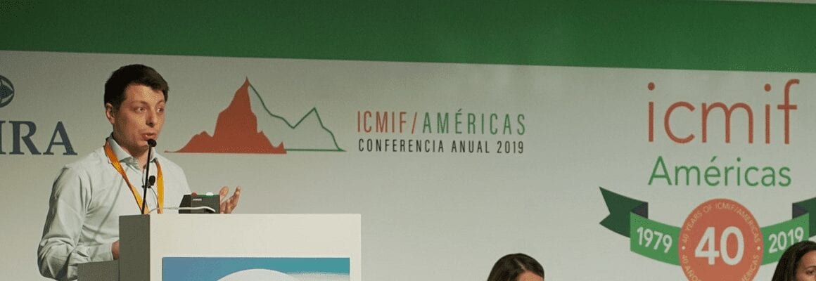 A man stands at a podium speaking into a microphone at the ICMIF/Americas Conferencia Anual 2019, with event banners and logos visible in the background.