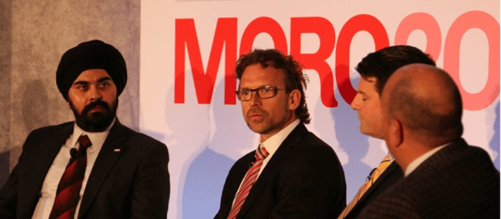 Four men in business attire sit and talk during a panel discussion in front of a sign with partially visible red text. One man wears a black turban, and another speaks while the others listen attentively.