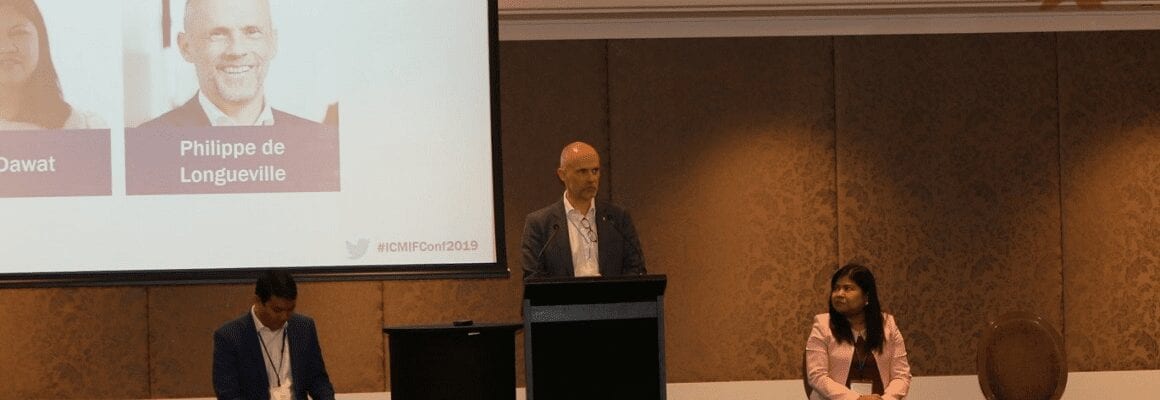 A man stands at a podium speaking at a conference. Two people sit on chairs on either side of him, and a large screen behind displays partial images and names. The setting appears to be professional and formal.