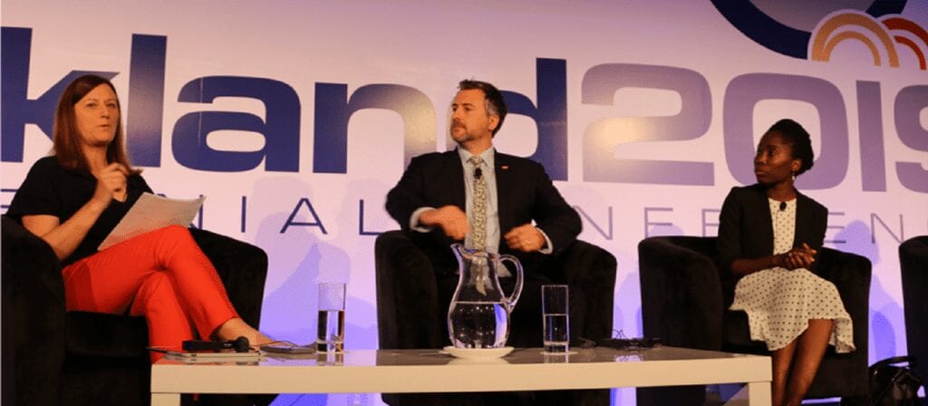 Three people sit on stage in armchairs, engaged in a panel discussion. A woman on the left holds papers, a man in a suit sits in the center, and a woman in a polka-dot dress sits on the right. Auckland 2019 is visible on the backdrop.