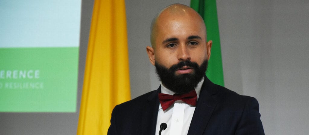 A man with a beard and bald head, wearing a dark suit, white shirt, and red bow tie, speaks at a podium. Yellow and green flags are visible in the background.