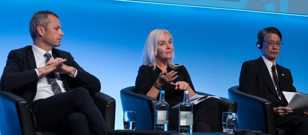 Three people in business attire sit on stage chairs against a blue background. The woman in the middle gestures while speaking, while the two men on either side listen, one with headphones. Water bottles and glasses are on the table in front.
