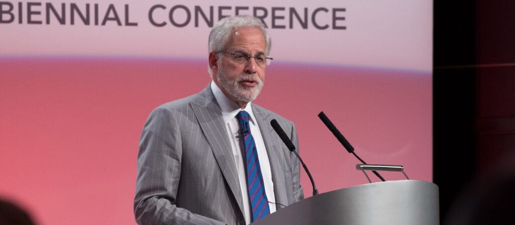 An older man with gray hair and glasses in a gray suit and striped tie speaks at a podium during a conference. The blurred background shows partial text reading BIENNIAL CONFERENCE.