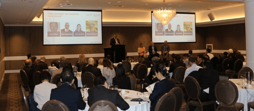 A conference room filled with seated attendees faces a stage where four panelists are seated, and one person stands at a podium. Two screens display images and panelist names for a discussion.