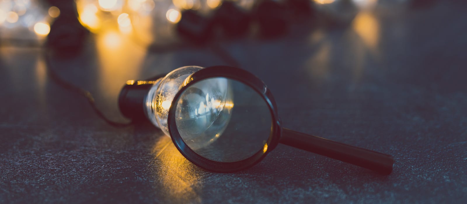 A magnifying glass rests on a table, magnifying part of a lightbulb lying nearby. Warm fairy lights are softly glowing in the blurred background, creating a cozy atmosphere.