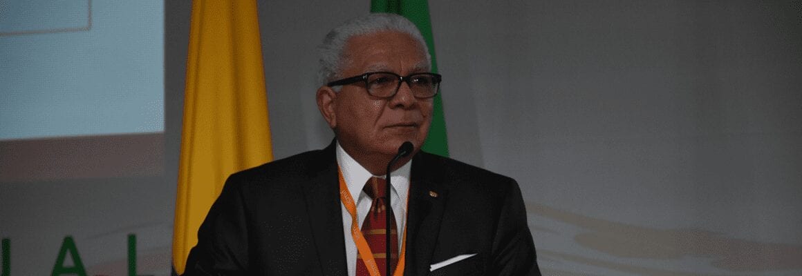 A man with gray hair and glasses, dressed in a suit and tie, speaks at a podium. A yellow and a green flag are visible in the background.