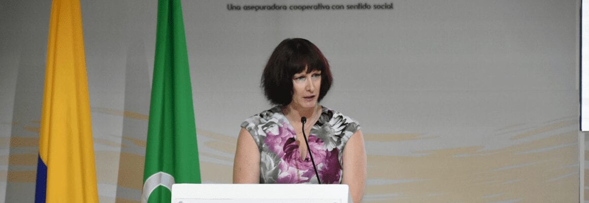 A woman with short brown hair wearing a floral dress speaks at a podium. Behind her are yellow and green flags and a backdrop with Spanish text.