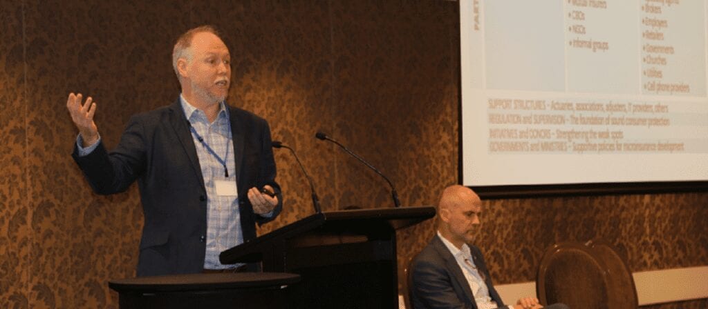 A man stands at a podium giving a presentation with a projected slide behind him, while another man sits nearby listening attentively. Both are dressed in business attire in a conference room.