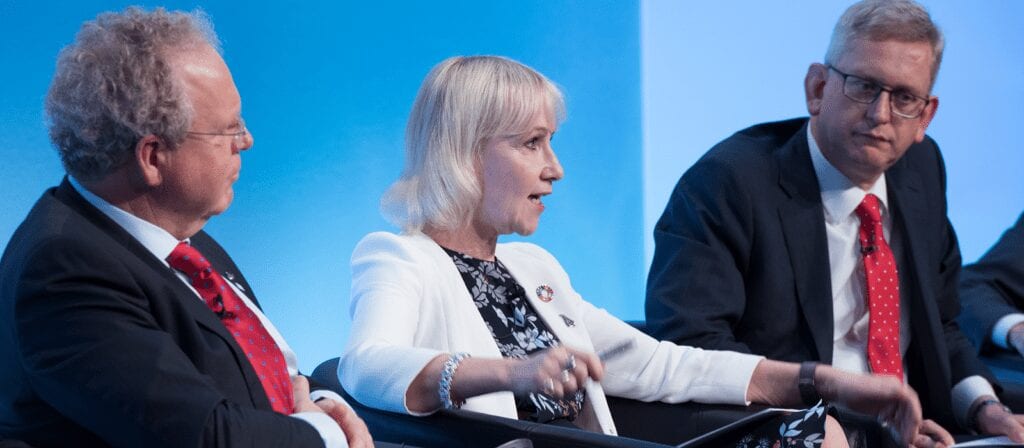 Three people in business attire are seated and engaged in a panel discussion against a blue background. The woman in the center is speaking, while the two men on either side listen attentively.