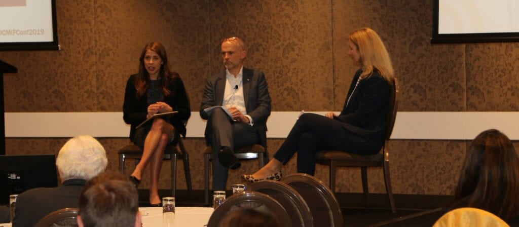 Three people in business attire sit on stage having a panel discussion at a conference, with an audience watching. Two women and one man are seated, the man is in the middle, and all appear to be engaged in conversation.