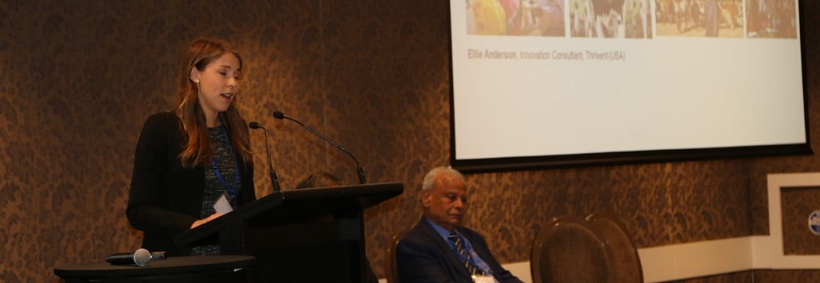 A woman stands at a podium speaking into a microphone while an older man sits beside her. A projection screen is visible behind them, displaying text and several small images.