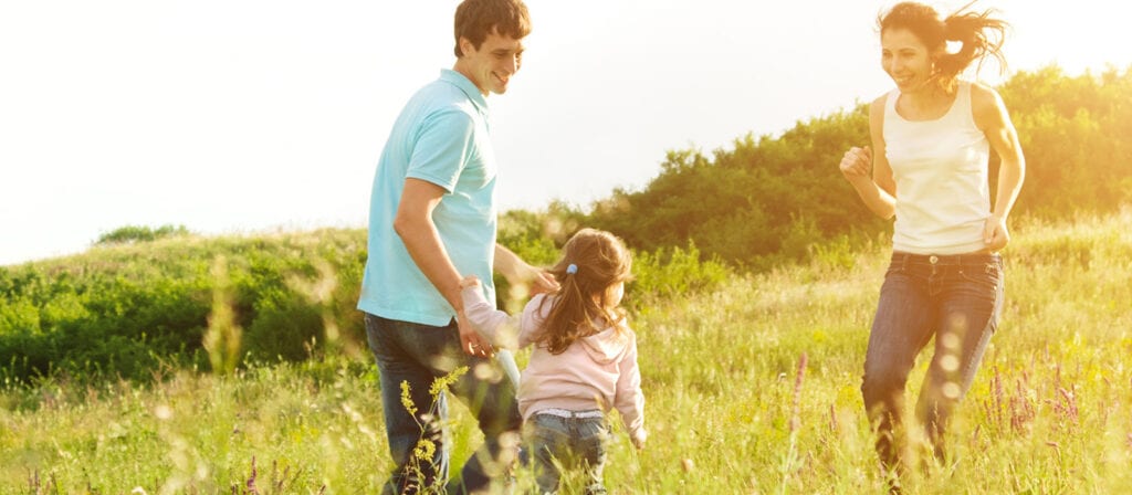 A man, woman, and young girl hold hands and run through a sunlit field with tall grass and wildflowers, smiling and enjoying the outdoors. Green hills and trees are in the background under a bright sky.