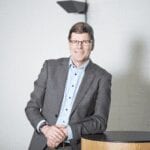 A man in a gray suit and glasses stands smiling, leaning on a round wooden table in a bright, modern room with a white wall and a black wall fixture in the background.