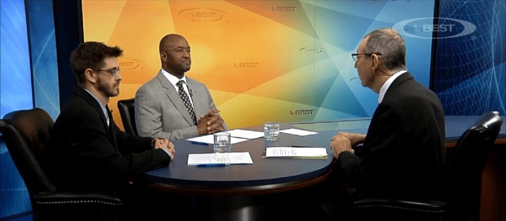 Three men in business attire sit around a round table having a discussion in a studio with colorful orange and blue backgrounds. Papers and glasses of water are on the table.