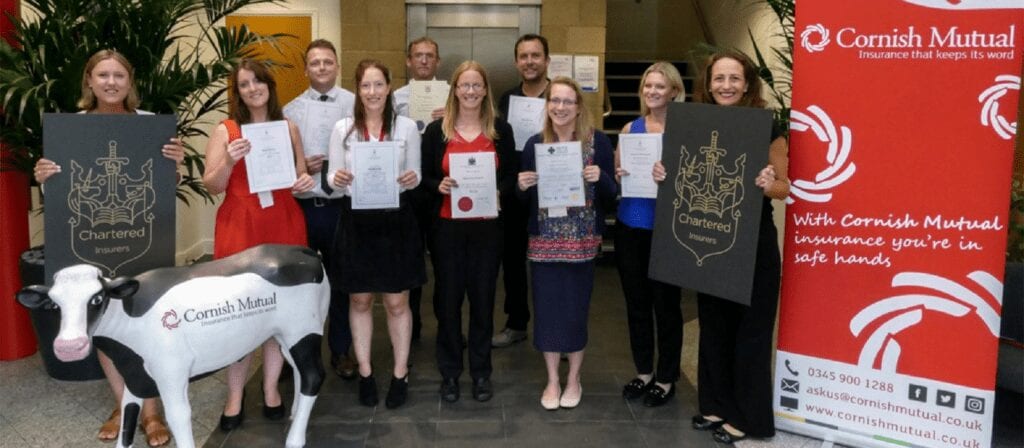A group of ten people stand indoors holding certificates, smiling at the camera. They are next to a cow statue and Cornish Mutual insurance banners. Two people hold large Chartered Insurer signs.