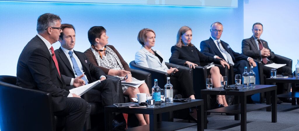 Seven professionally dressed individuals sit on a stage panel, engaged in discussion. They are seated in black chairs with microphones and water bottles on the tables in front of them, against a blue-lit background.