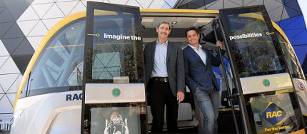 Two men in business attire smile while standing at the open doors of a modern, yellow-and-white autonomous vehicle with Imagine the possibilities written above them. Geometric building panels are visible in the background.