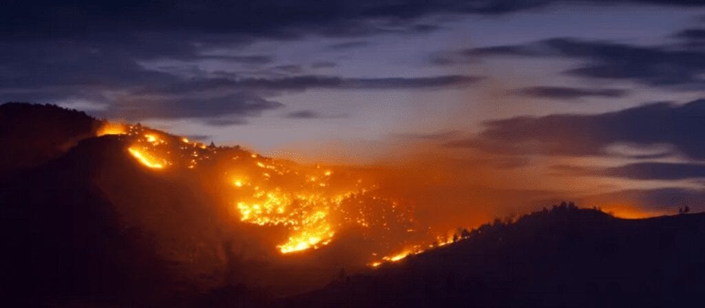 A wildfire spreads across a hillside at dusk, with bright orange flames illuminating the dark landscape and thick smoke rising into the evening sky.