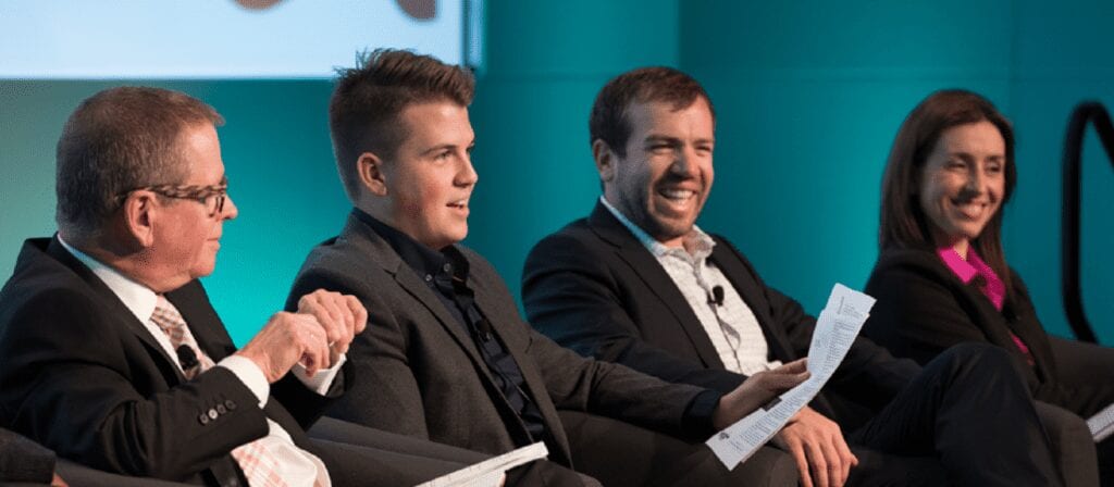 Four people in business attire sit on a panel, holding papers and smiling while engaged in discussion against a teal background.