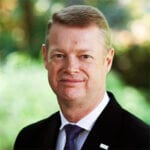 A middle-aged man with short light brown hair, wearing a black suit, white shirt, and purple tie, stands outdoors in front of a blurred green background. He is smiling slightly.