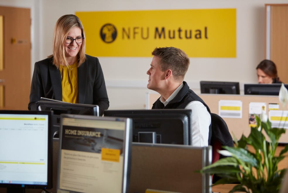 Two office workers have a conversation at their desks in a modern office. The NFU Mutual logo is visible on a yellow sign in the background, and computer monitors display insurance-related content.