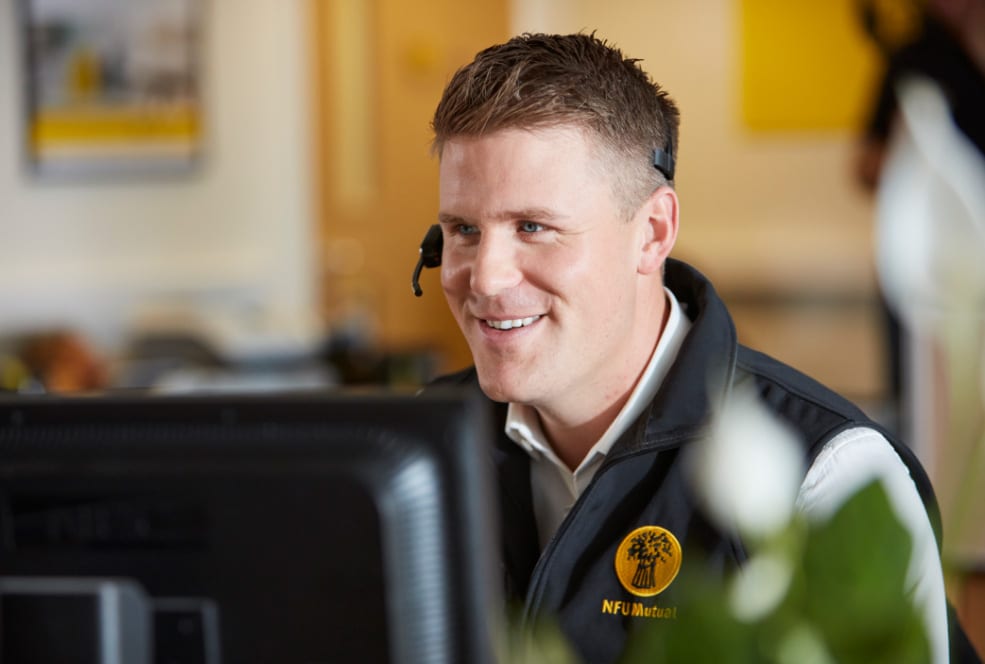 A smiling man wearing a headset and an NFU Mutual vest sits at a desk, looking at a computer screen in a bright office environment.