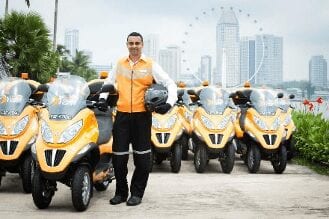 A man in an orange and white uniform stands holding a helmet next to a row of identical yellow motor scooters, with a city skyline and Ferris wheel in the background.