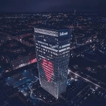 A tall building at night displays a large red heart made of lights on its facade, with the cityscape illuminated in the background. The building has Polsat written on top.