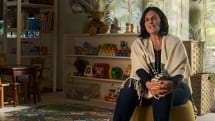 A woman with dark hair sits on a yellow ottoman in a cozy, well-lit room filled with childrens toys, books, and colorful decorations, with shelves and small chairs in the background.
