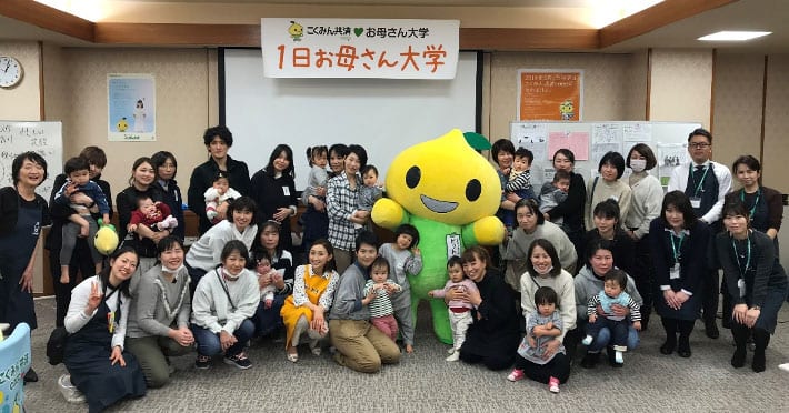 A large group of adults and young children pose together in a room, smiling at the camera. In the center stands a person in a bright yellow mascot costume. Japanese text banners hang on the wall behind them.