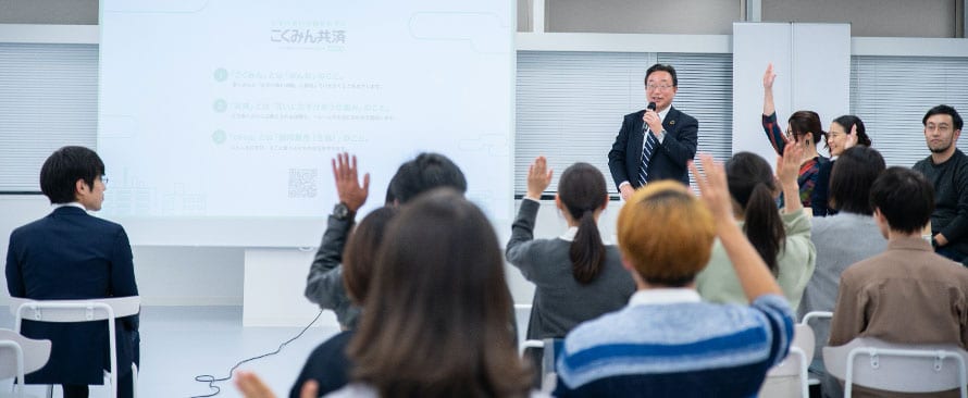 A man in a suit speaks at a podium in front of a projection screen, while several audience members, seated in rows, raise their hands to participate. Other presenters stand nearby, and the setting appears to be a seminar or workshop.