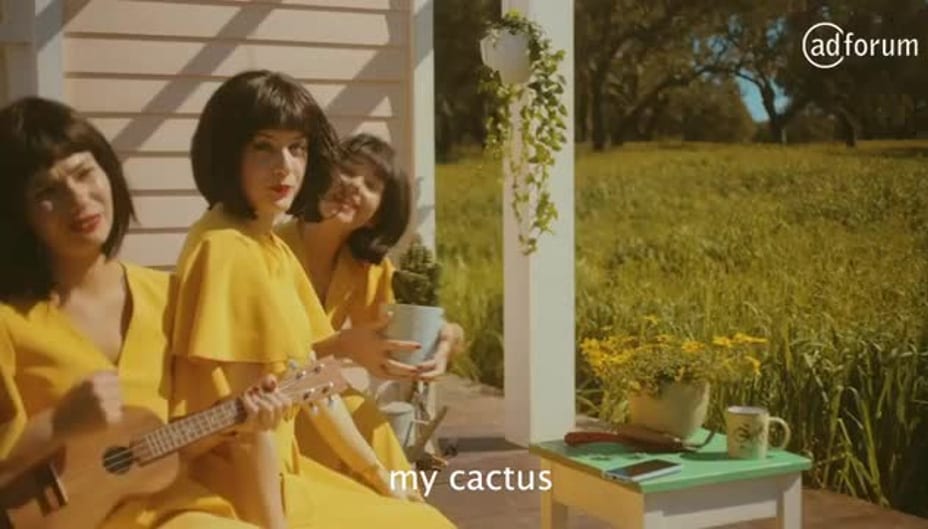 Three women in yellow dresses with short black wigs sit on a porch. One holds a ukulele, another holds a potted cactus. A green table with flowers and a phone is nearby. The subtitle reads my cactus.
