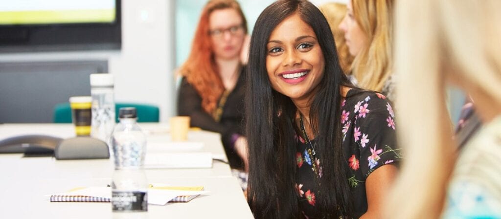 A woman with long dark hair and a floral dress smiles while sitting at a conference table with others, notebooks, and water bottles in a brightly lit meeting room.