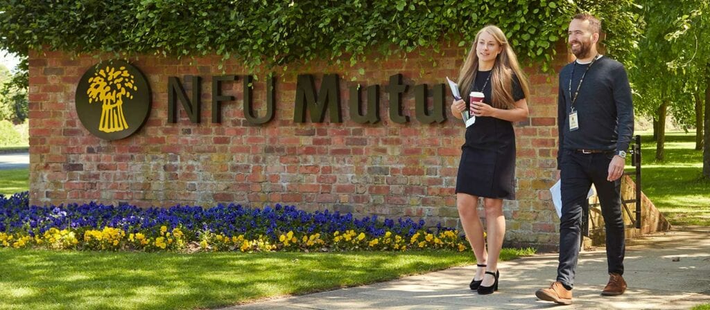Two people, a woman and a man, walk and talk outside an NFU Mutual office building, holding drinks. They are on a sidewalk beside a brick wall with the company logo and name, surrounded by flowers and greenery.