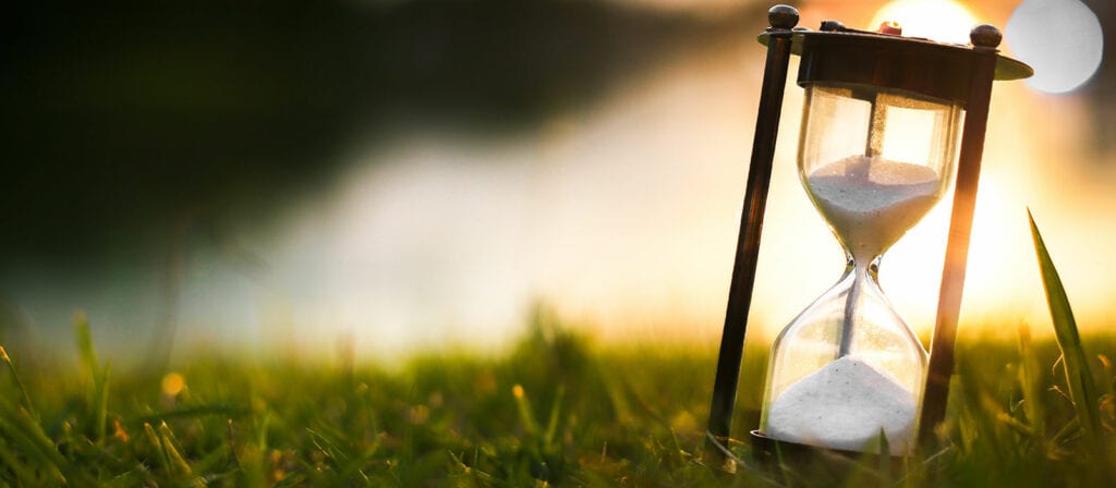 A close-up of an hourglass with white sand, placed on green grass outdoors, with sunlight creating a warm, blurred background.
