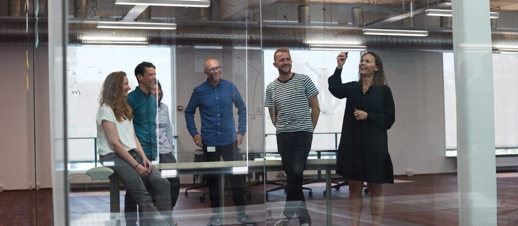 Five people stand in a modern office, gathered around a glass wall with writing on it. One woman writes on the glass while the others watch and smile, suggesting a collaborative meeting or brainstorming session.