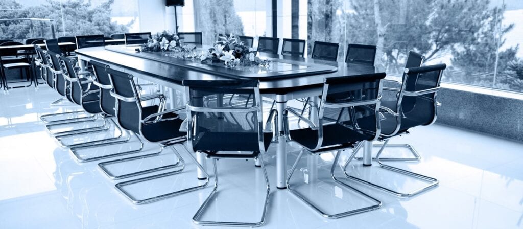 A modern conference room with a large rectangular table surrounded by black mesh chairs. The table is decorated with flower arrangements, and large windows provide a view of trees outside.