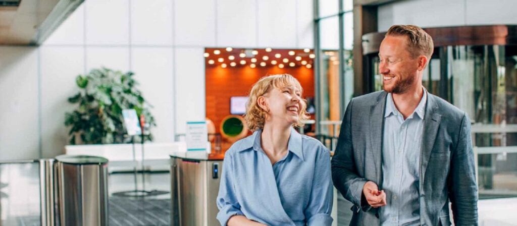A woman and a man dressed in business attire walk and laugh together in a modern office lobby with glass walls, plants, and warm lighting in the background.